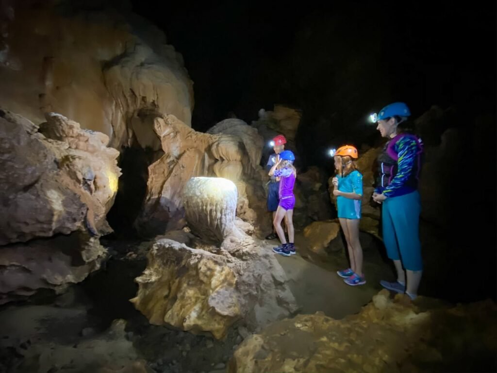 People wearing headlamps examine cave formations while exploring an underground cavern. Natural rock structures are illuminated against a dark, secluded background.