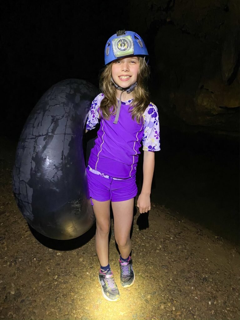 A person wearing purple hiking gear and a helmet stands next to a large rock in a dark cave. A headlamp illuminates the path in this underground scenic view.