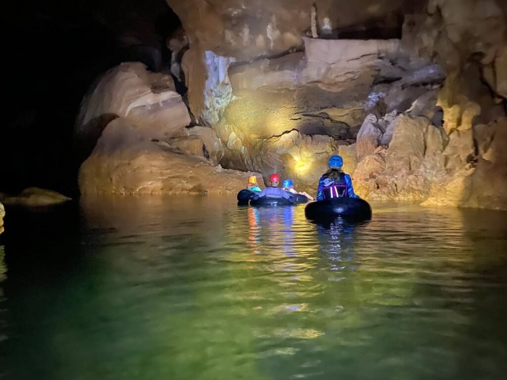 People tubing through a cave with illuminated rock walls and calm green water.