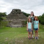 Belize Day 7 A small group stands before a historic stone temple structure surrounded by lush greenery and overcast skies. The setting features ancient architecture, steps, and tropical foliage.