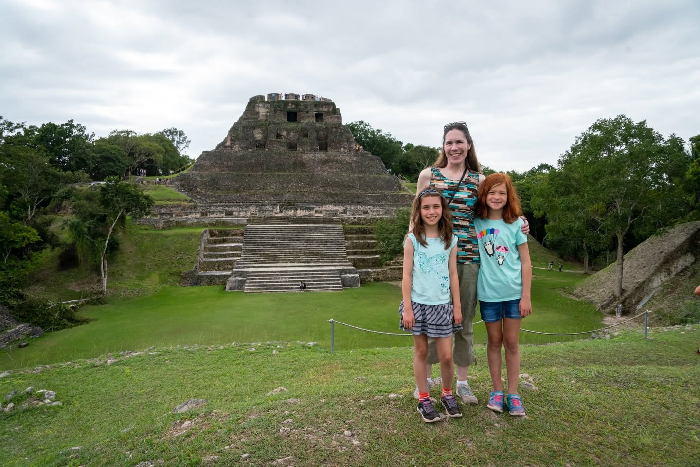 Belize Day 8 A small group stands before a historic stone temple structure surrounded by lush greenery and overcast skies. The setting features ancient architecture, steps, and tropical foliage.