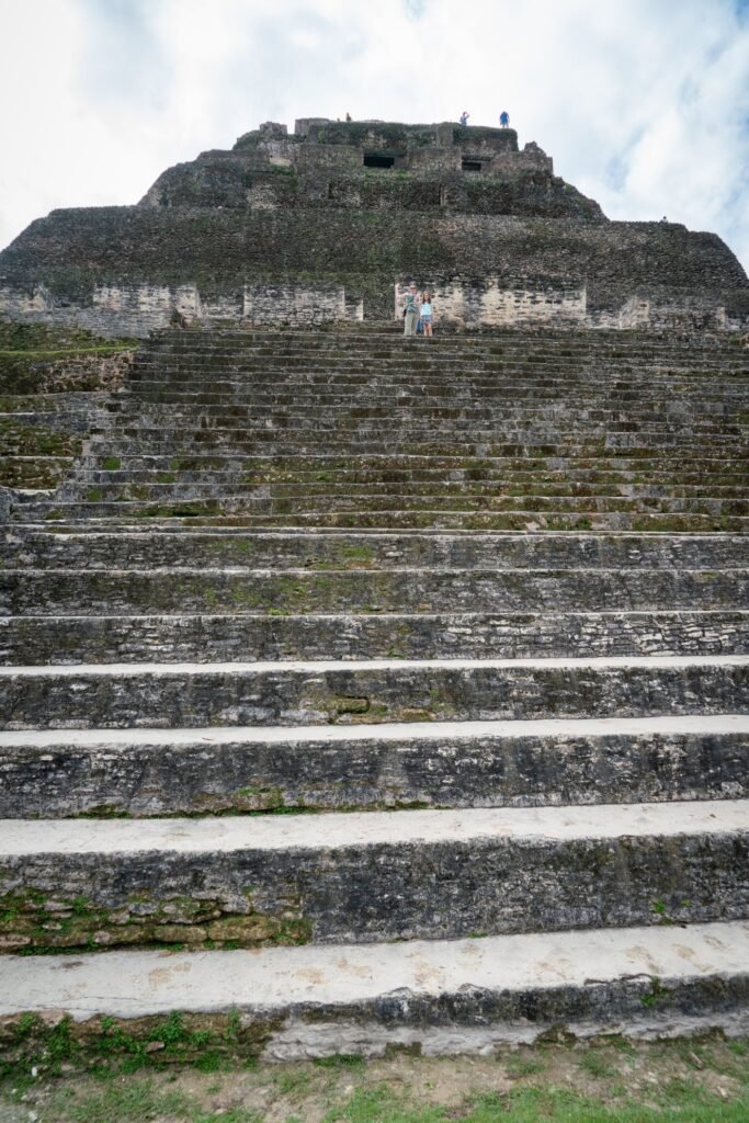 Closely layered stone steps leading up to a lush, overgrown Mayan pyramid. The ancient structure stands tall against a soft cloudy sky in Mexico.