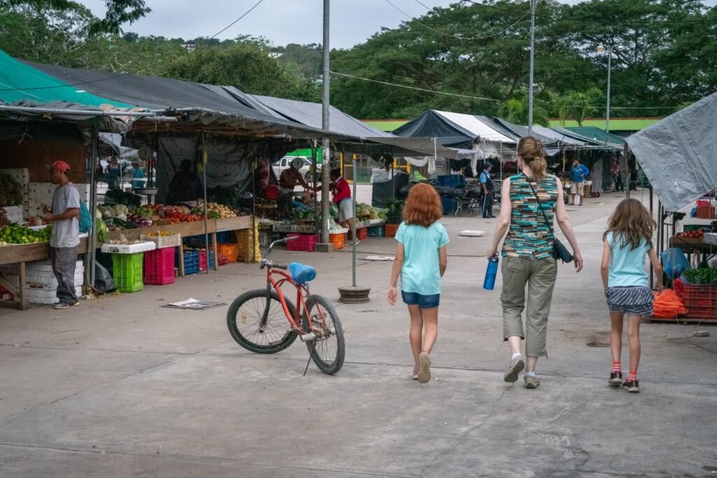 People walking through an open-air market surrounded by stalls with produce and a bicycle.
