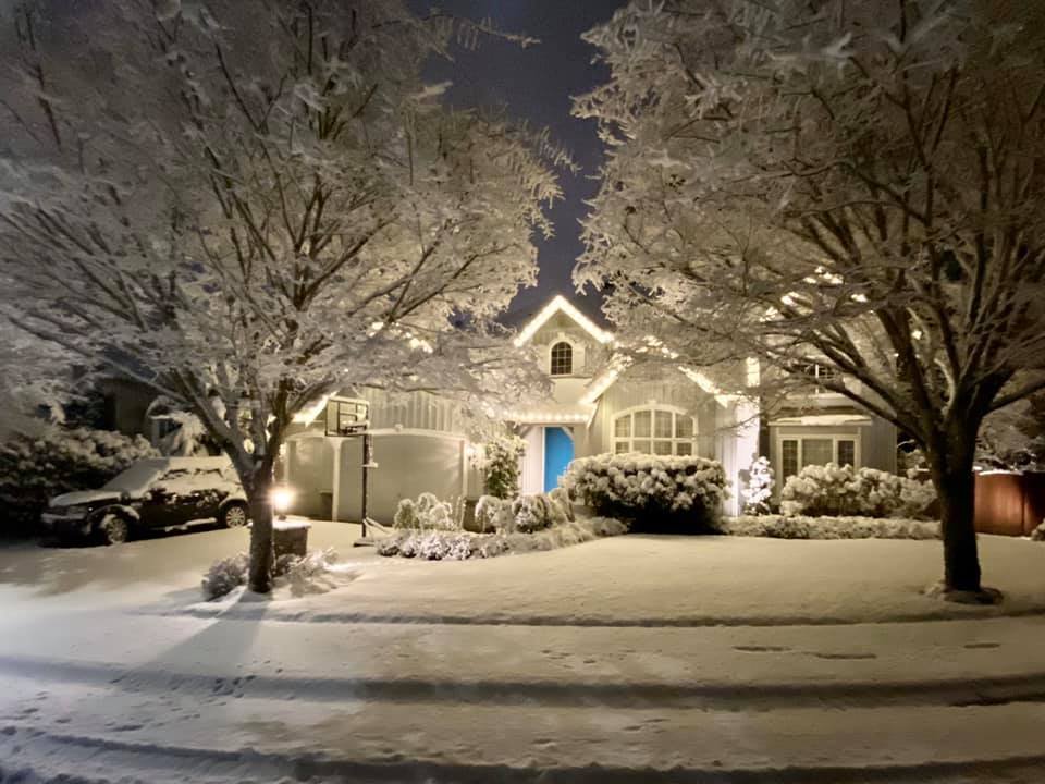 Residential home covered in snow at night with glowing lights and snow-covered trees in the foreground.