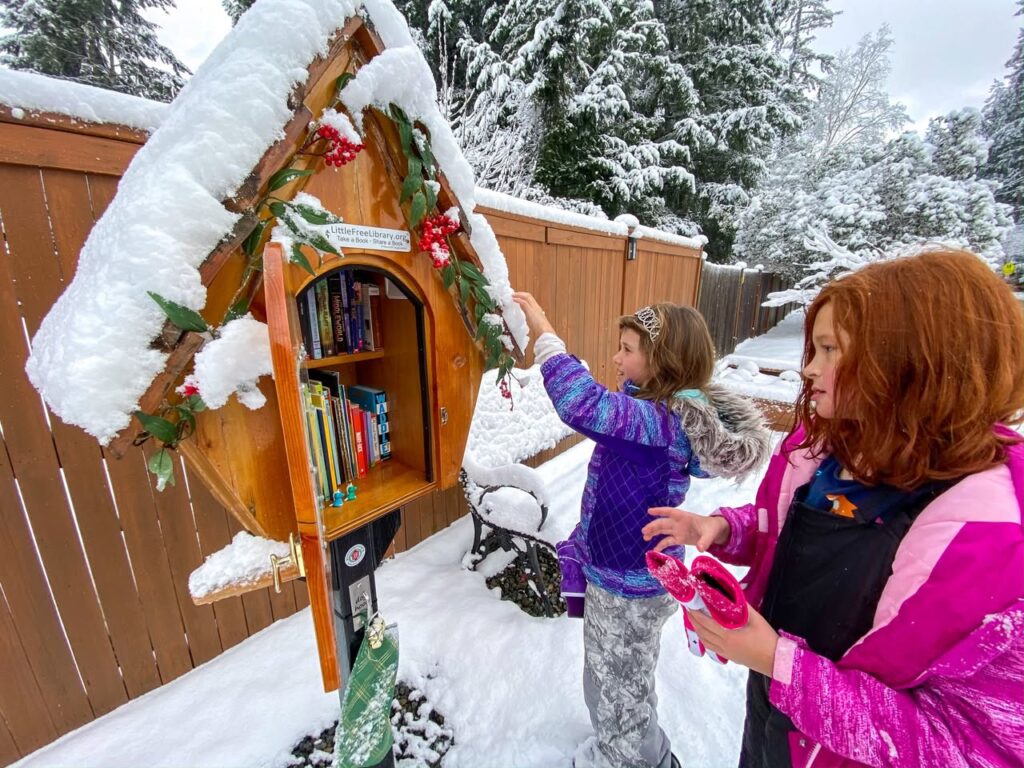 A snowy Little Free Library decorated with greenery in a Pacific Northwest backyard. A child reaches for a book while another person watches nearby.