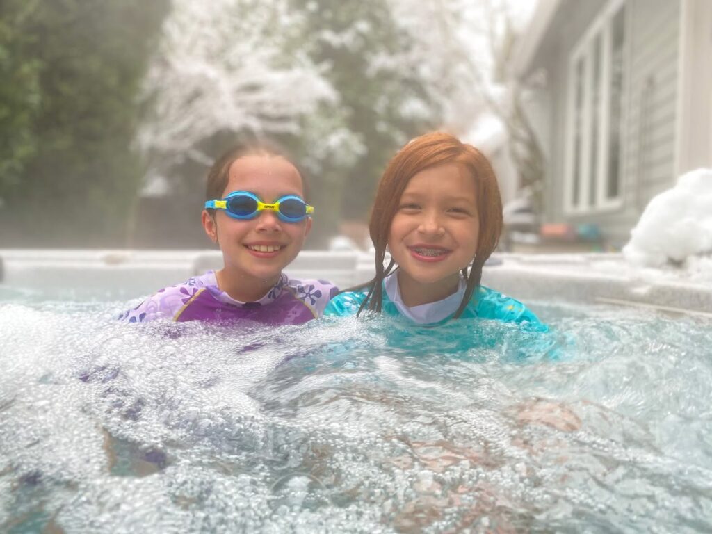 Two smiling children enjoy a hot tub on a snowy day in the Pacific Northwest. Steam rises from the bubbling water against a backdrop of a snow-covered house and trees.