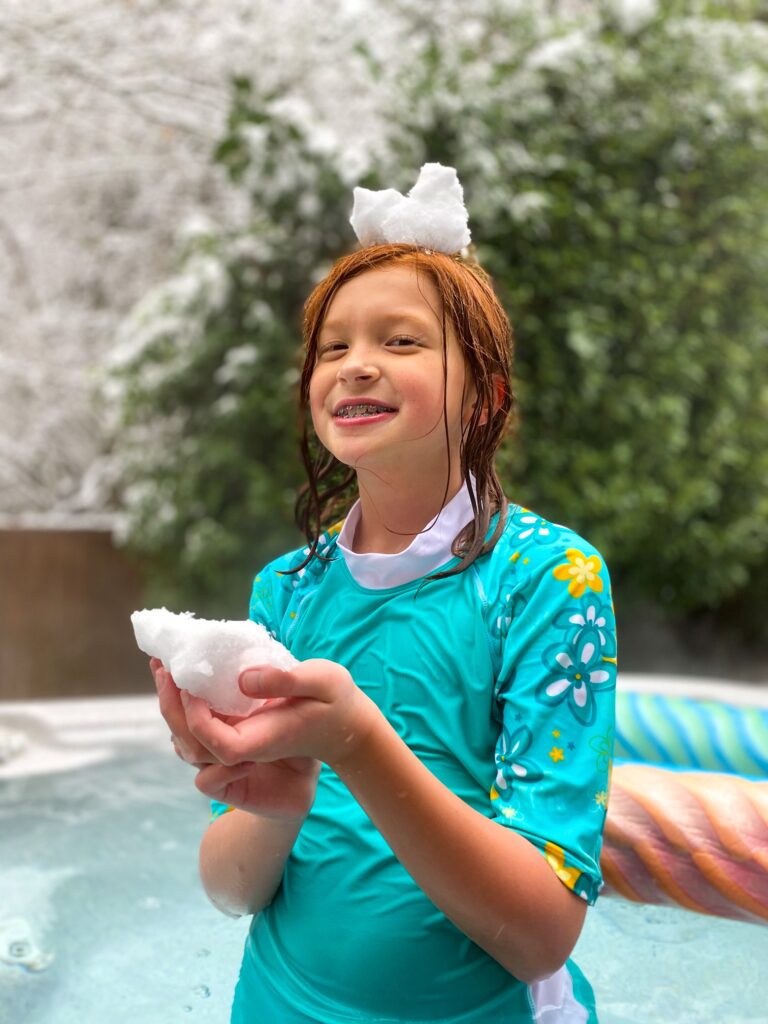 A young person in a blue rash guard enjoys holding snow during a snowy day at an outdoor hot spring.