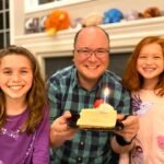 A smiling family holds a slice of cake with a lit candle in their cozy living room, celebrating a special moment together.