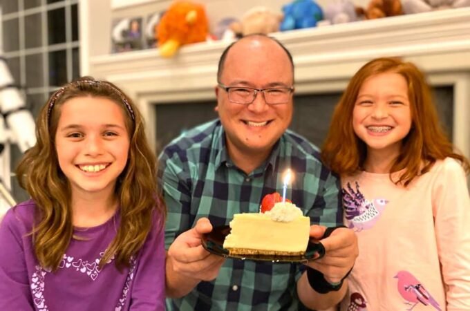A smiling family holds a slice of cake with a lit candle in their cozy living room, celebrating a special moment together.