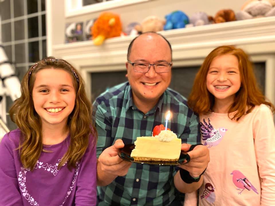 A smiling family holds a slice of cake with a lit candle in their cozy living room, celebrating a special moment together.