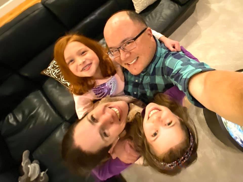 A family of four takes a cheerful selfie together on a black leather couch.