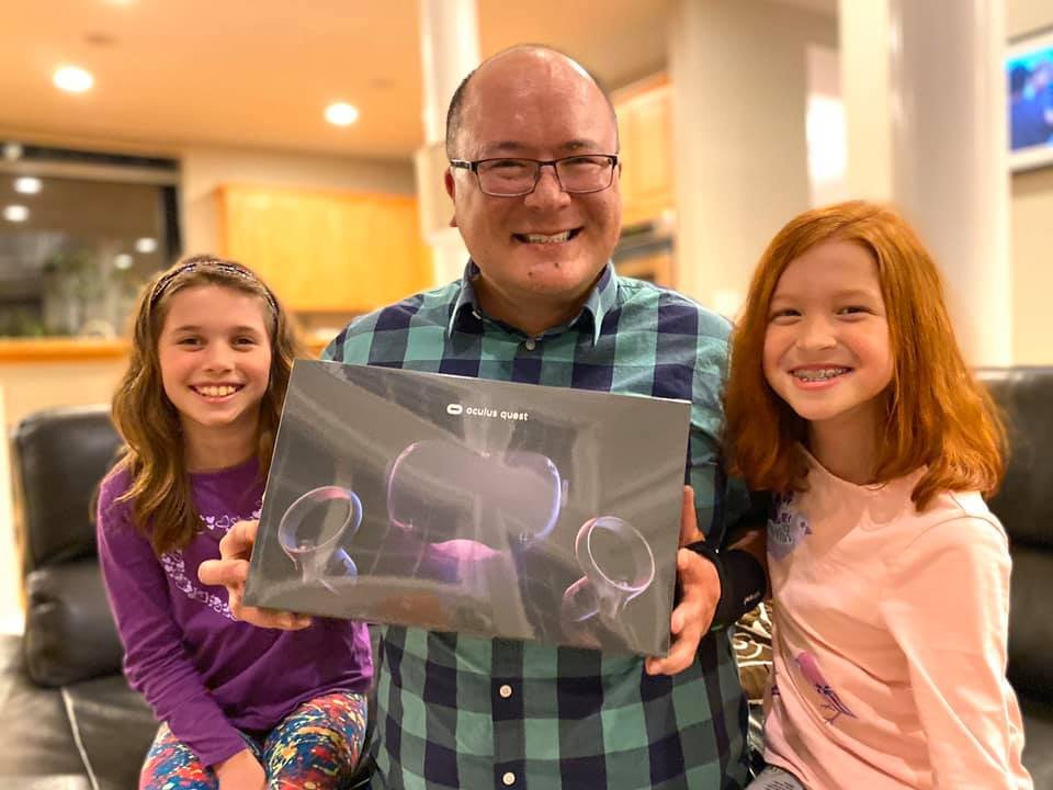 A photo of a family holding an ultrasound image in a warm indoor environment. The group, including a man and two girls, is smiling as they display something special together.