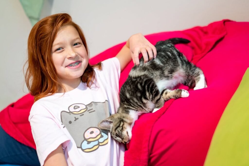 A red-haired girl smiles warmly while gently petting a tabby cat resting on a bright pink blanket.