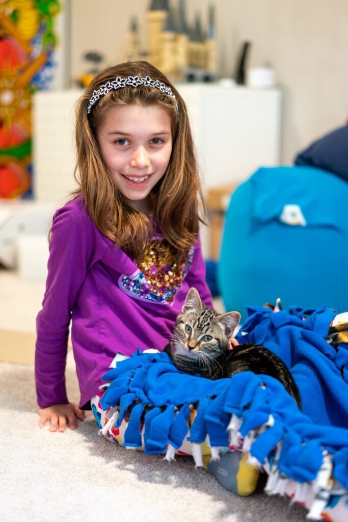 A girl in a purple shirt smiles while sitting on the floor next to a cat resting on a blue and white knitted blanket. The scene captures a warm, homey atmosphere in a Pacific Northwest living room.