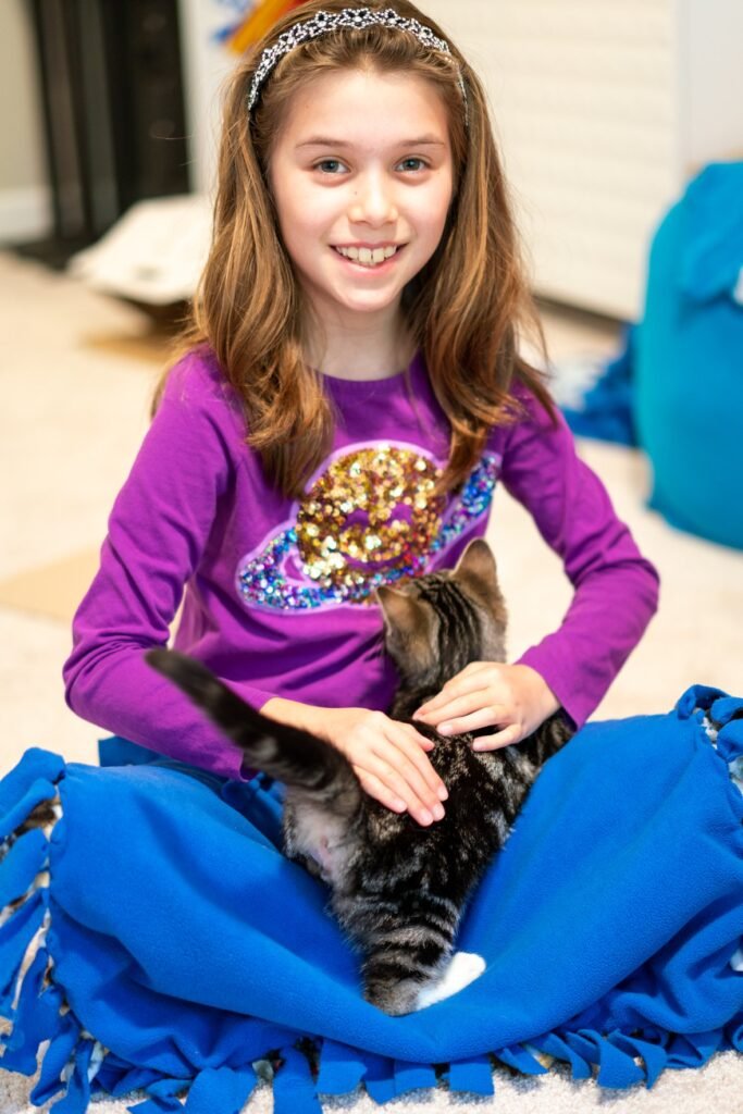 A young girl in a purple shirt sits on a blue fringed blanket, gently holding and petting a tabby kitten.
