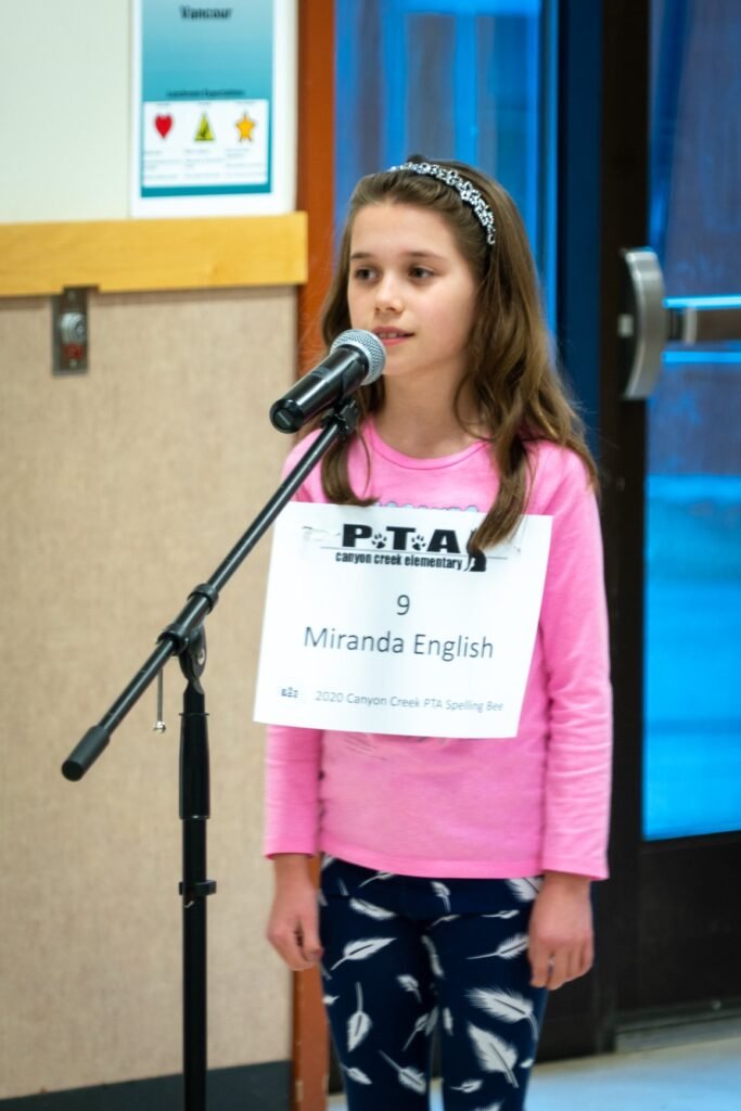 A girl in a pink shirt stands at a microphone with a PTA sign during a school event.