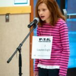A child standing at a microphone with a PTA sign showing their name. Wearing a pink and black striped shirt.