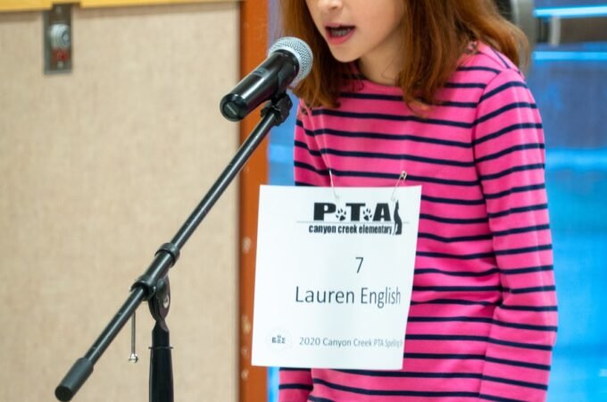 A child standing at a microphone with a PTA sign showing their name. Wearing a pink and black striped shirt.