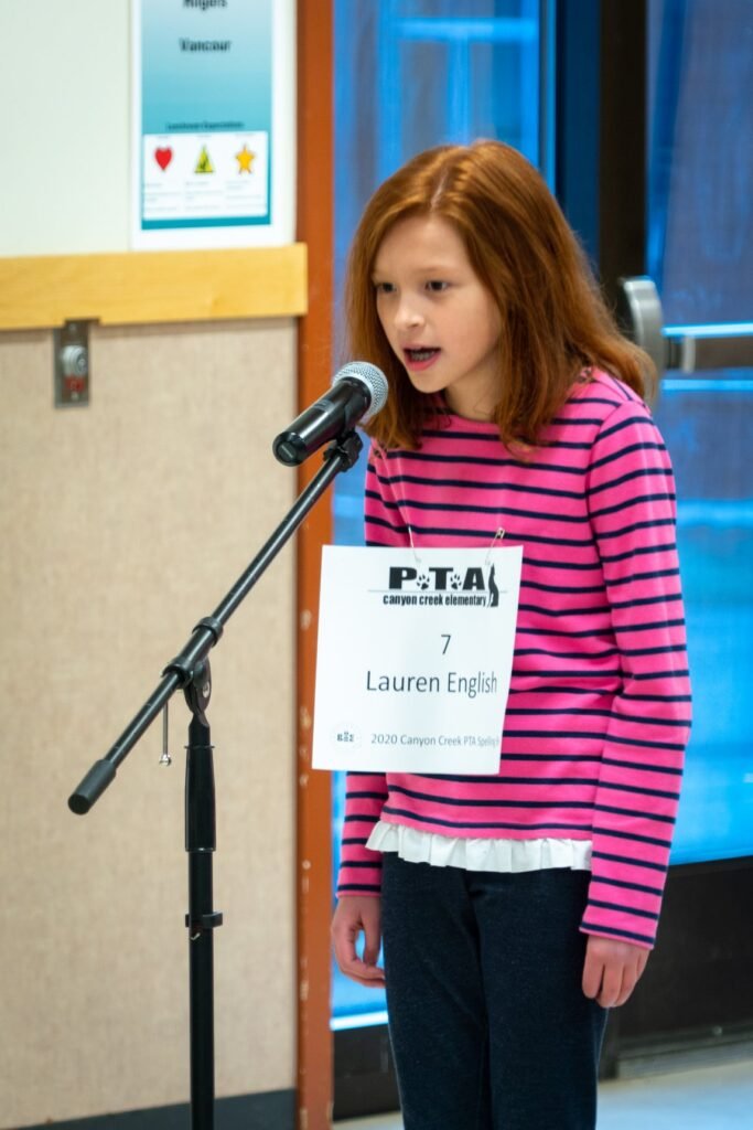 A child standing at a microphone with a PTA sign showing their name. Wearing a pink and black striped shirt.