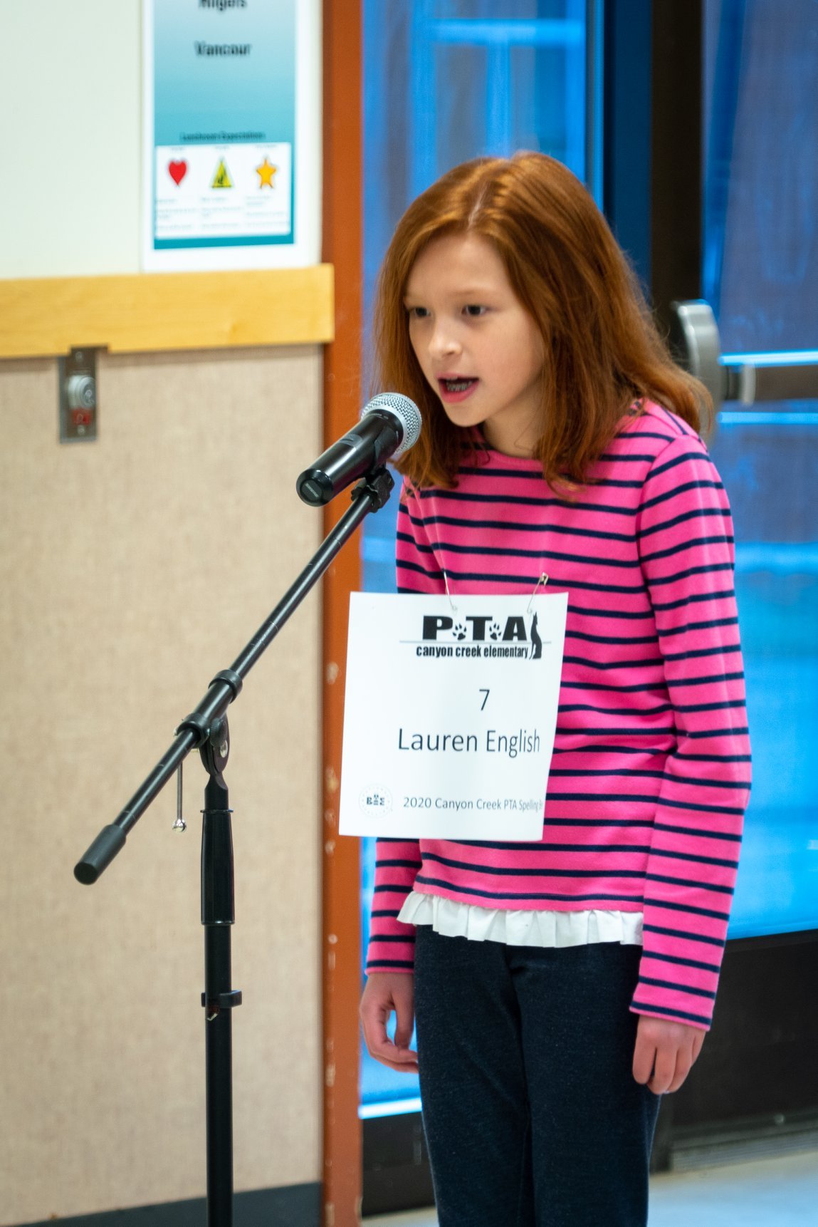 Spelling Bee A child standing at a microphone with a PTA sign showing their name. Wearing a pink and black striped shirt.