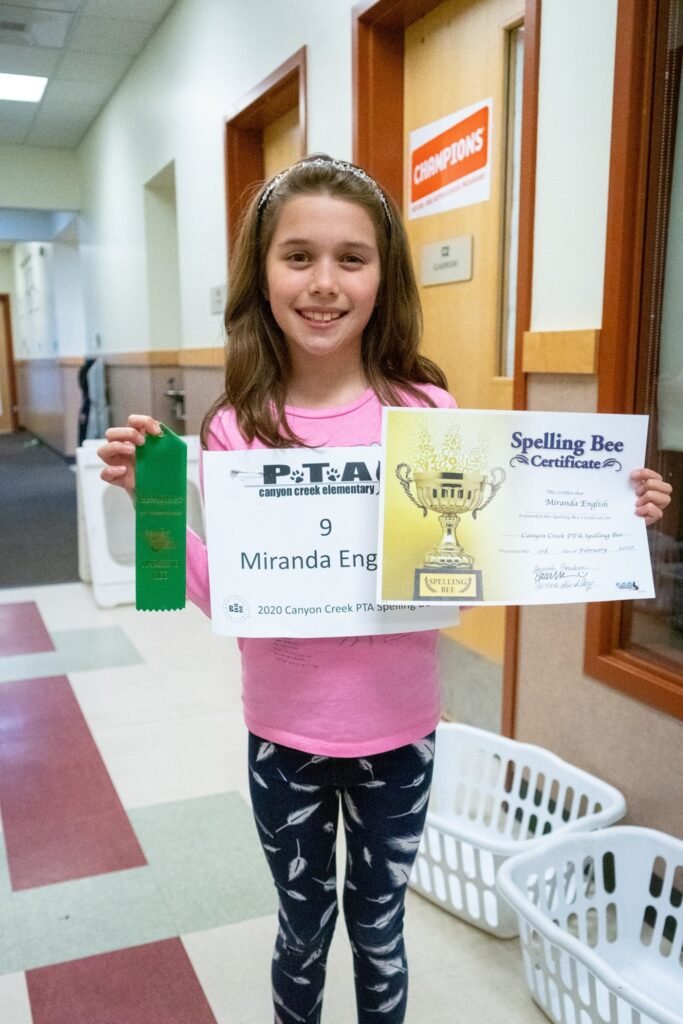 A child holds a Spelling Bee certificate and green trophy in a school hallway.