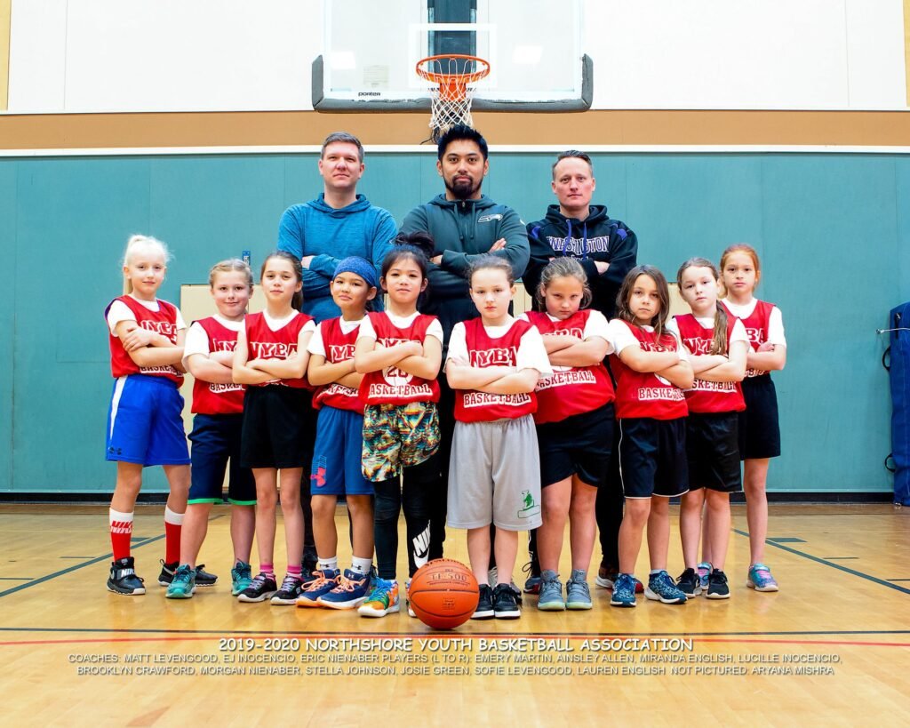 A youth basketball team and coaches posing together in a gym with a basketball hoop visible in the background.