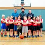 A youth basketball team and coaches posing together in a gym with a basketball hoop visible in the background.