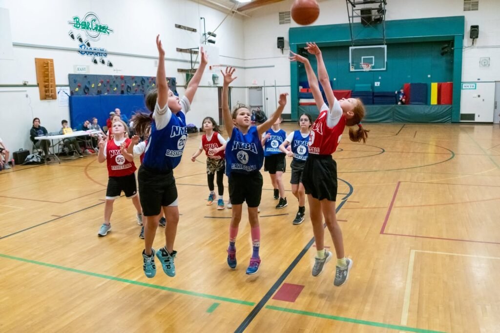 Young athletes in red and blue jerseys leap for a basketball during an indoor game.