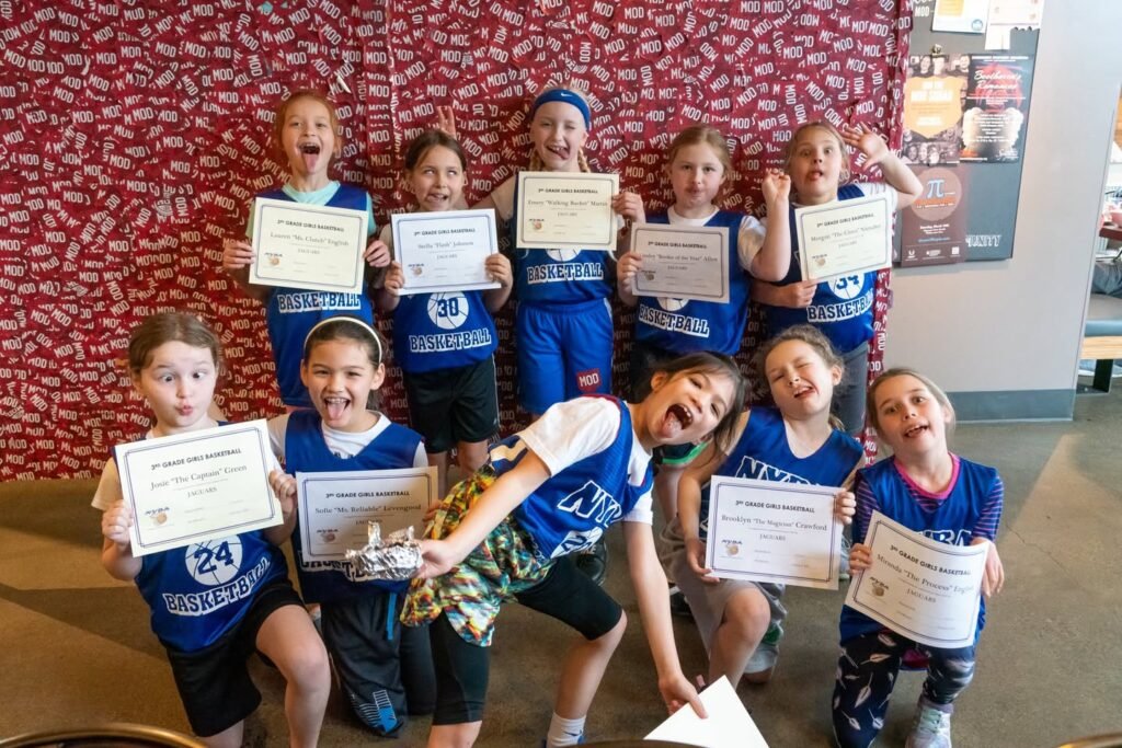 Children in blue swim team uniforms holding certificates during a group photo. The team celebrates their achievements in the pool facility.