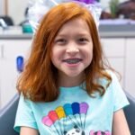 A smiling person with vibrant red hair and braces wearing a teal t-shirt with a panda parachute design.