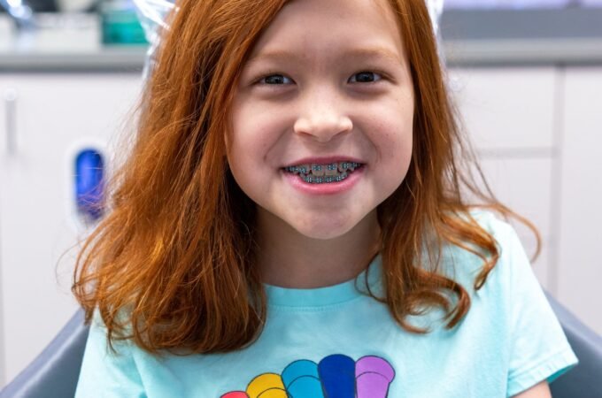 A smiling person with vibrant red hair and braces wearing a teal t-shirt with a panda parachute design.