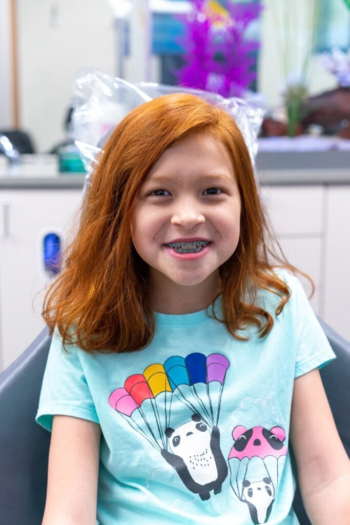 A smiling person with vibrant red hair and braces wearing a teal t-shirt with a panda parachute design.