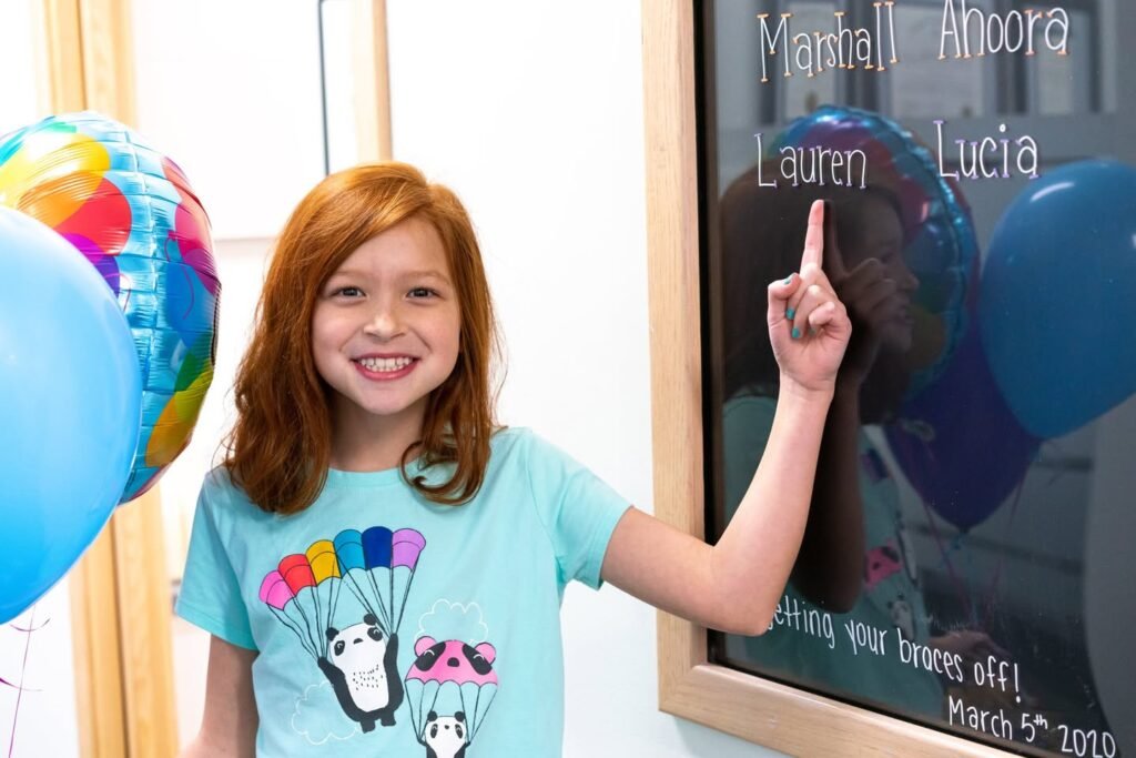 A smiling red-haired child in a teal panda shirt points to names written on glass next to colorful balloons.