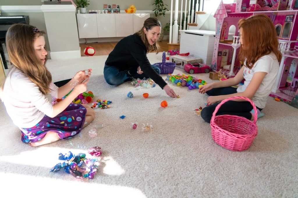 A woman and two children gathered on a carpeted floor collecting colorful plastic eggs and a pink basket during an indoor Easter activity.
