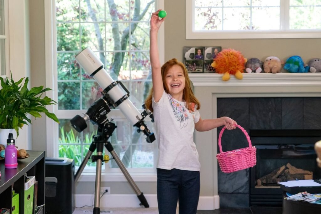 A person stands near a tripod-mounted telescope, holding a pink basket and a green egg. A fireplace with plants on the mantel is in the background.