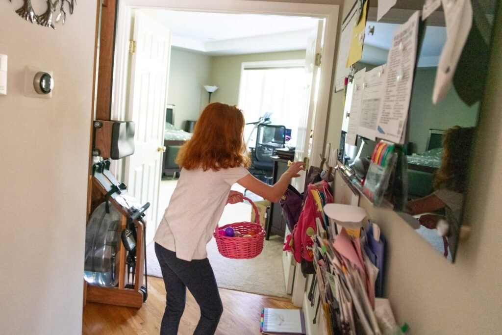 A person in a white shirt and dark pants reaches into an open shelving unit in a home hallway. A red basket and mirror are visible in the background.