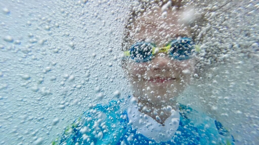 A child wearing goggles splashes into a pool. The photo is taken from underwater capturing the face and blue shirt.