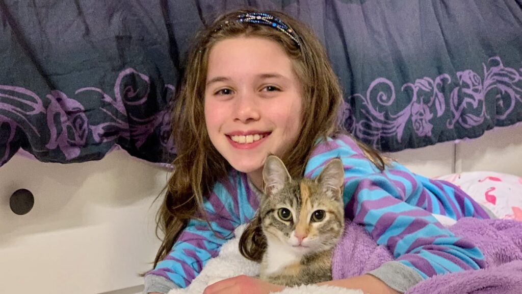 A young girl smiles while holding a tabby cat in her bed, sharing a cozy moment under a purple blanket.