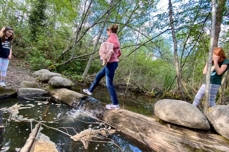 People crossing a small creek via a log in a lush forest setting.