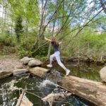 A nature enthusiast carefully crosses a log bridge over a clear stream. A dense forest of birch trees surrounds the path in the Pacific Northwest.