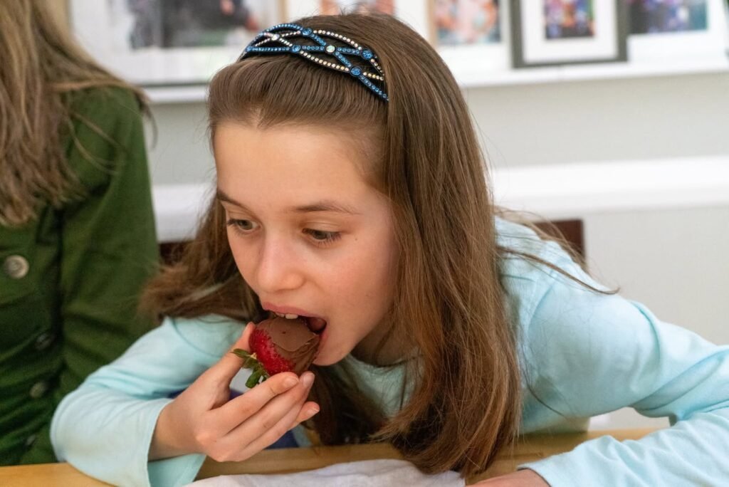 A young girl wearing a blue top and a headband bites into a chocolate-covered strawberry in an indoor setting.