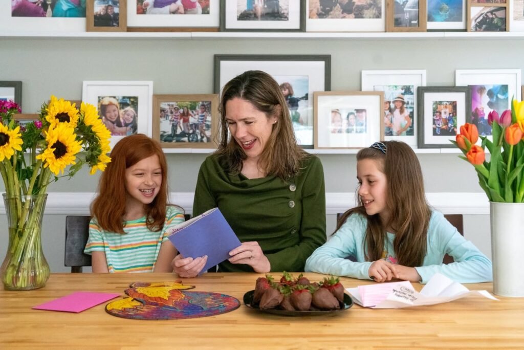 A smiling adult and two children share a moment at a table with sunflowers and tulips. Framed photos decorate the shelves behind them in a cozy home setting.