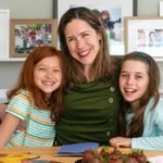 Smiling woman and two girls seated at a dining table with framed family photos displayed on shelves behind them.