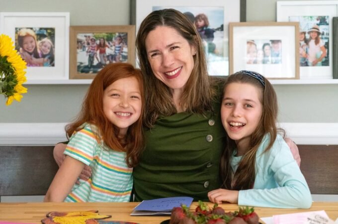 Smiling woman and two girls seated at a dining table with framed family photos displayed on shelves behind them.