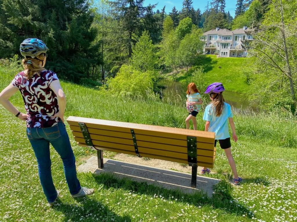 A cyclist rests on a wooden bench in a grassy, flower-filled park with a scenic view.