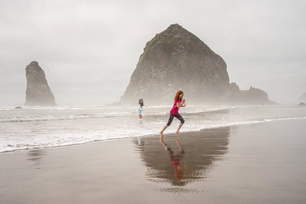 Two people running on a wet beach with rock formations and fog in the background. A person in a red shirt running in front, another in a white shirt behind.