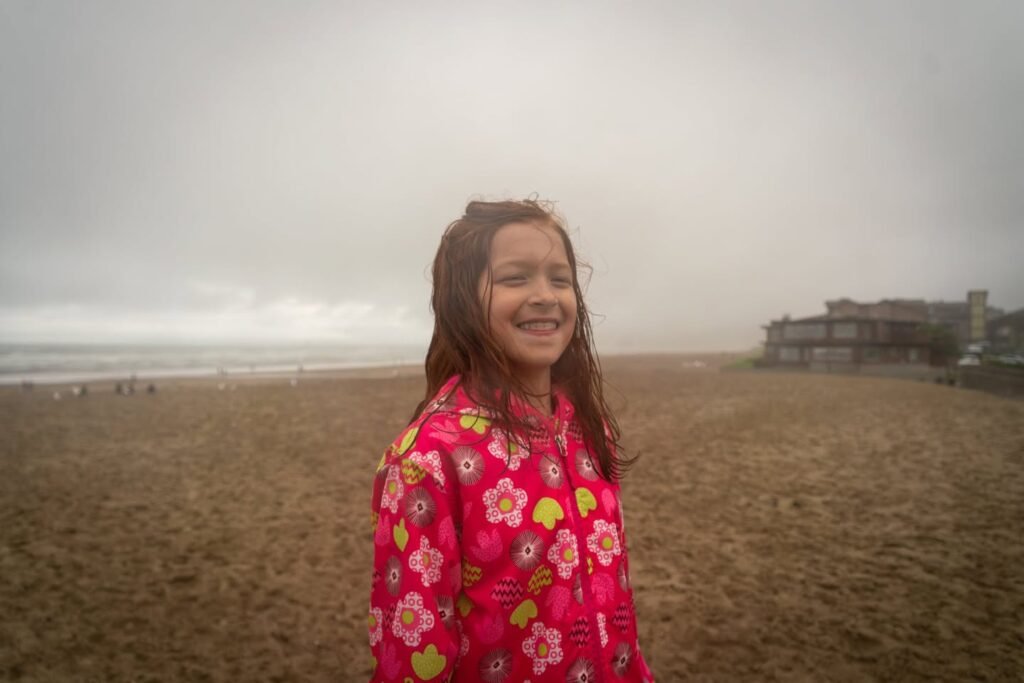 A person in a floral jacket stands on a foggy beach. A distant building can be seen through the hazy mist.