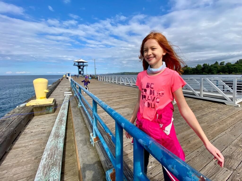 A young person smiles while leaning on a blue railing of a wooden pier extending into the water under a partly cloudy blue sky.