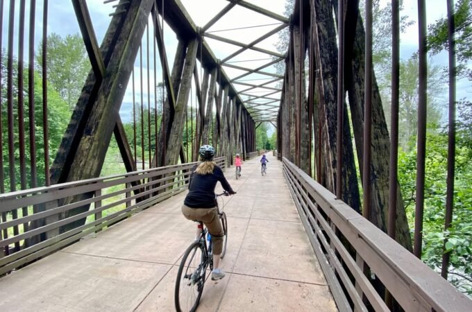 A cyclist rides across a historic iron truss bridge surrounded by forest. The view looks down from behind a railing.