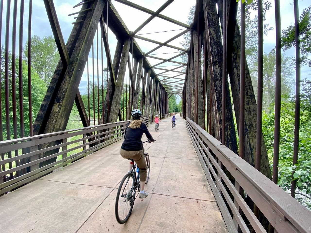 Sequim to Port Angeles Bike Ride A cyclist rides across a historic iron truss bridge surrounded by forest. The view looks down from behind a railing.